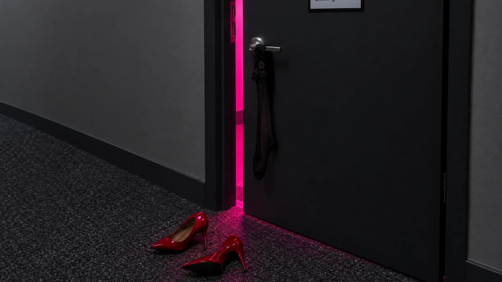 A dark door marked Dressing Room 1 stands ajar, pink light spilling out over red heels and a stocking.