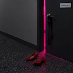 A dark door marked Dressing Room 1 stands ajar, pink light spilling out over red heels and a stocking.