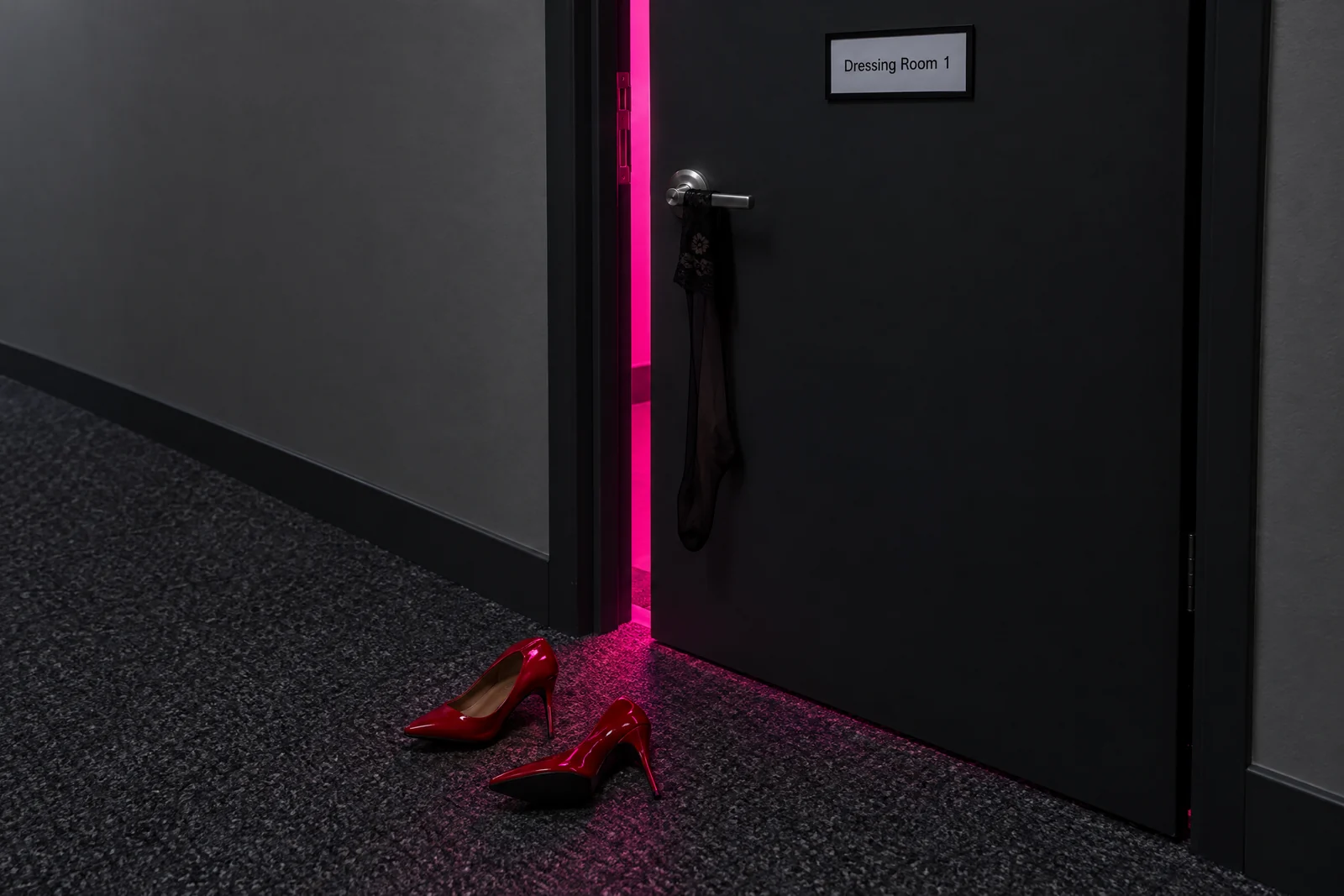 A dark door marked Dressing Room 1 stands ajar, pink light spilling out over red heels and a stocking.